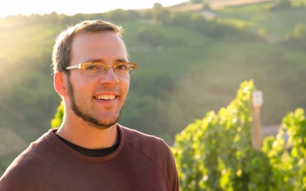 Mann mit Brille lächelt im Sonnenlicht vor grüner Hügellandschaft und Weinreben