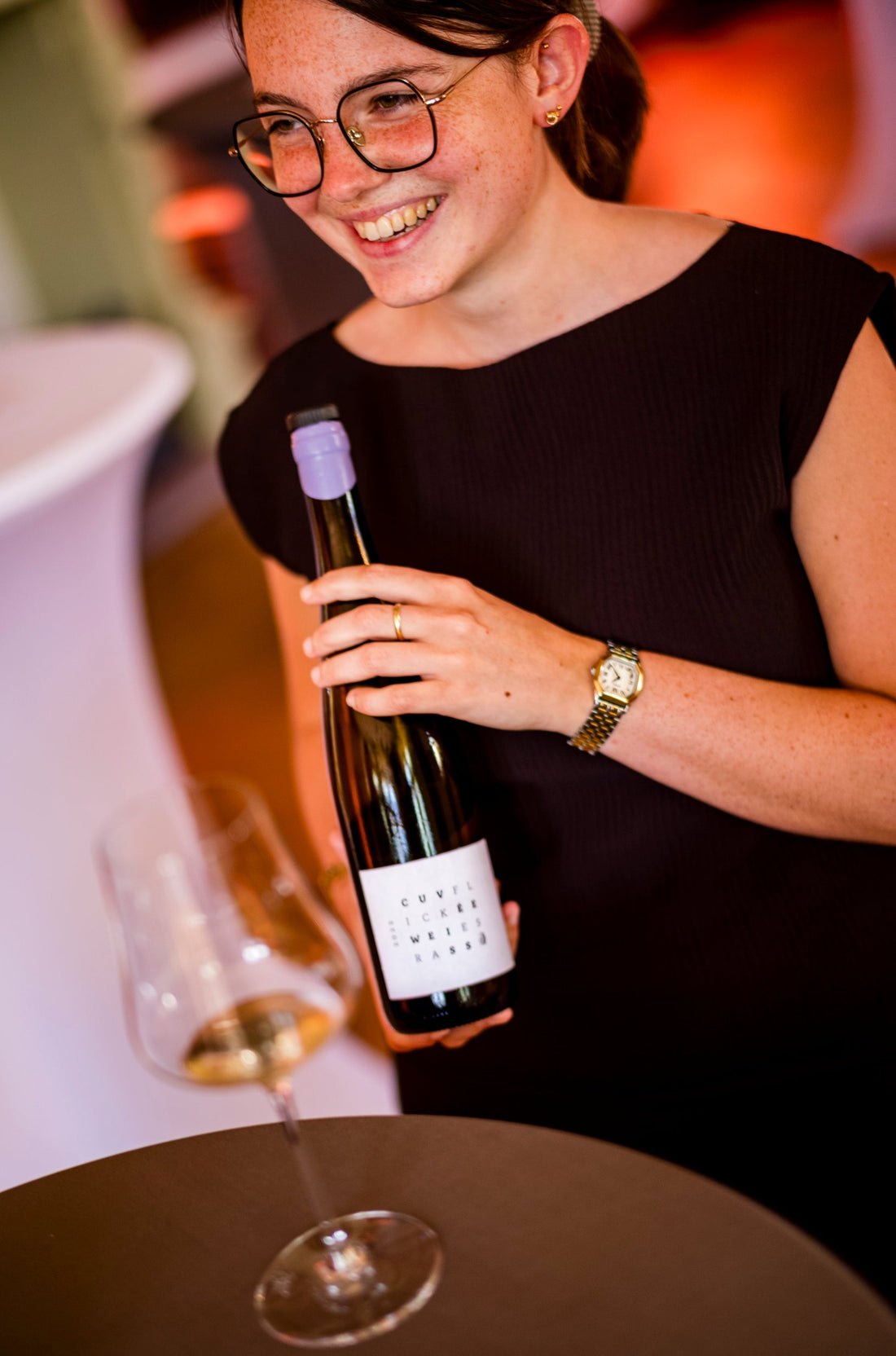 Frau mit Brille hält Weinflasche und lacht, Weinglas auf Tisch, Weindegustation Event