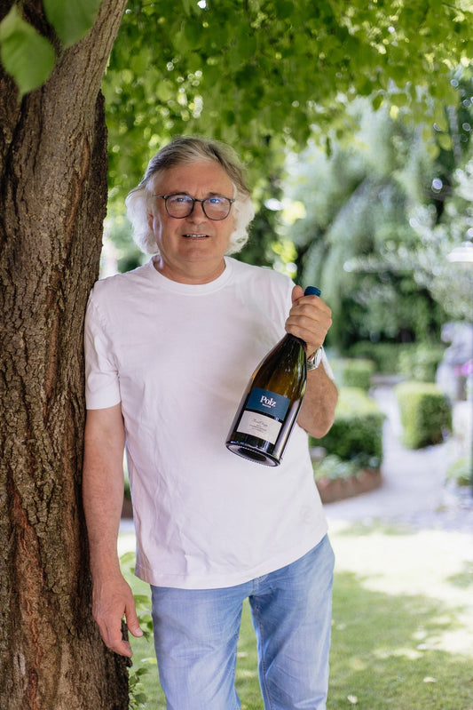 Mann mit weißem T-Shirt hält Polz Chardonnay Weinflasche im Garten neben Baum